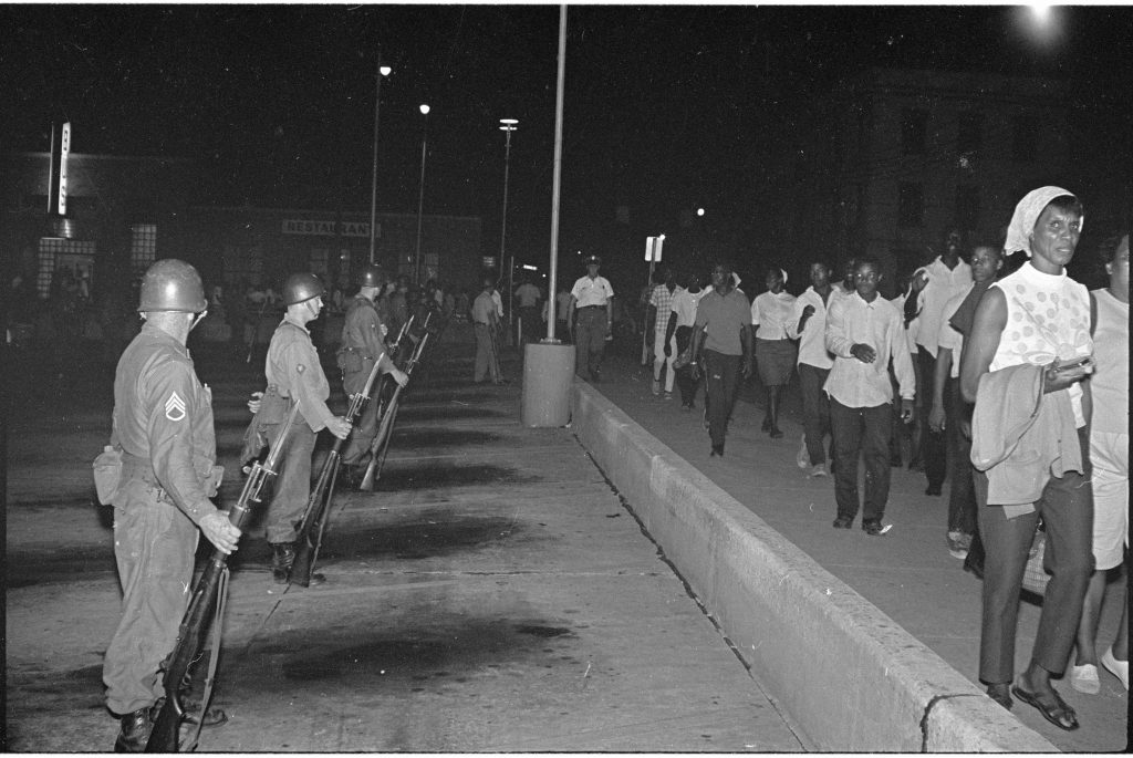 Black and white photograph of a group of protesters watched by armed guards