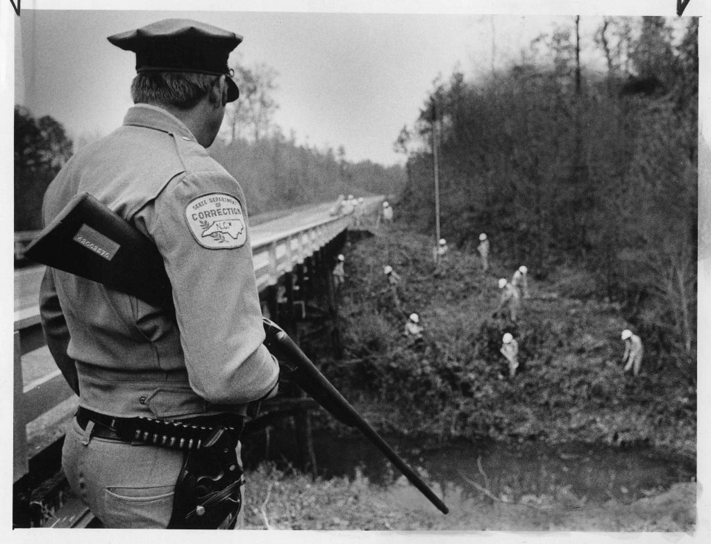 Black and white photograph of an armed guard standing by a bridge watching a work crew on the bank below