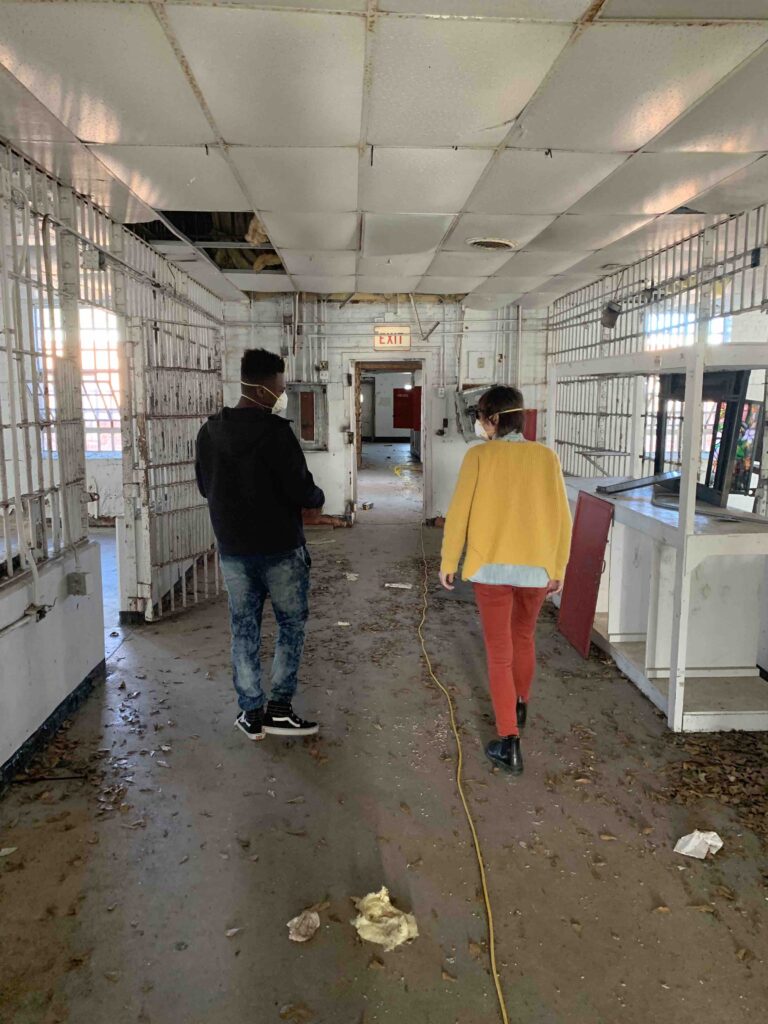 Two people walk through the interior of a decommissioned prison, with broken ceiling tiles and debris on the floor