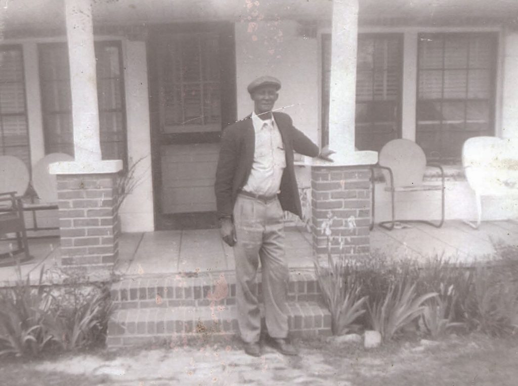 Black and white photograph of a Black man in a suit and hat standing in front of a house's front porch
