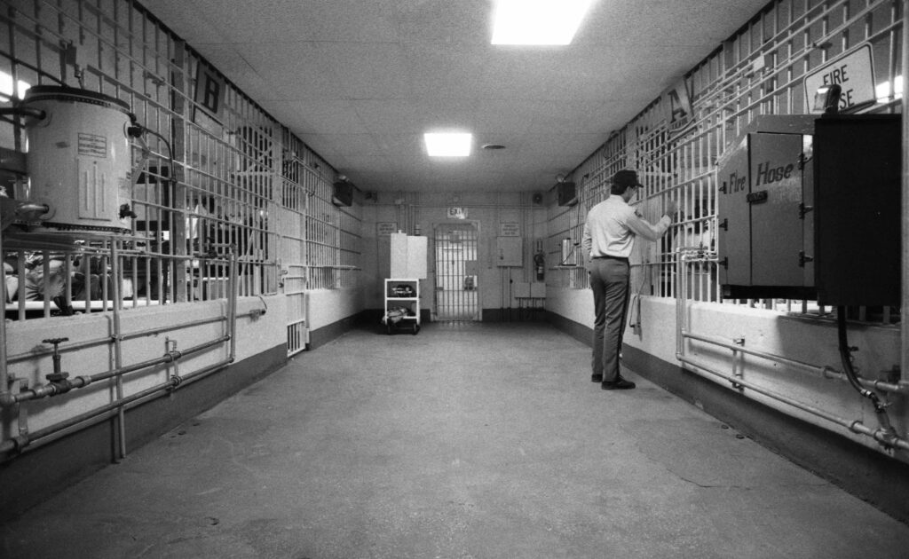 Black and white photograph of prison corridor. A guard is looking into prison cells. 