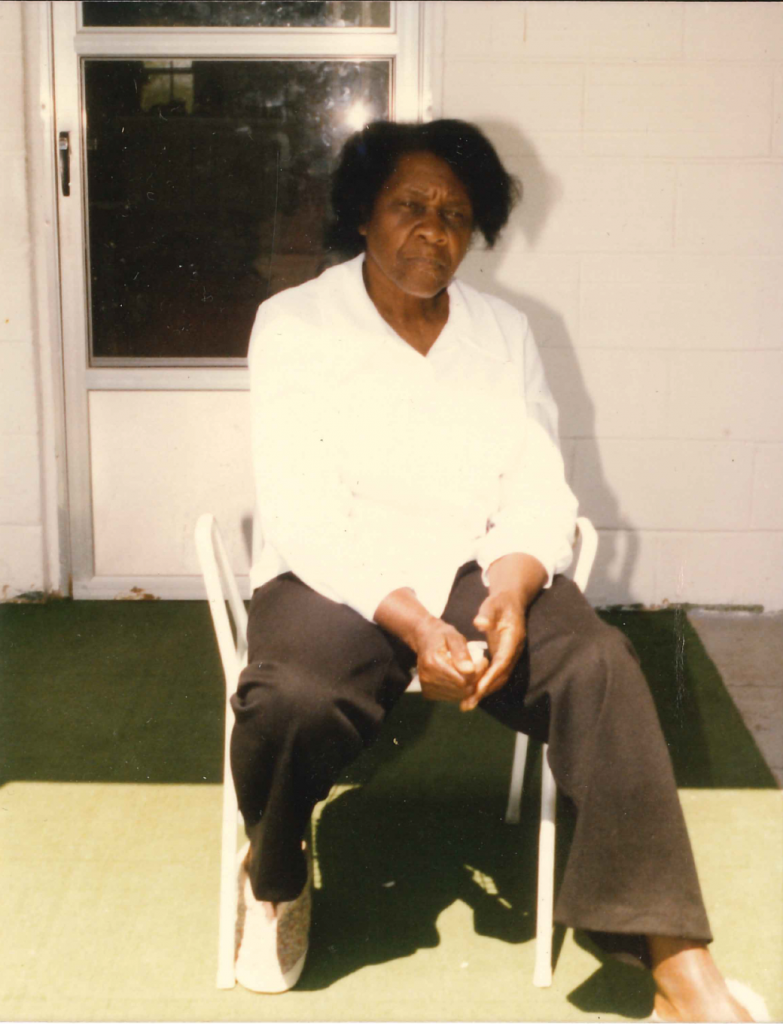 Color photograph of a Black woman in a chair in front of a cinder block wall