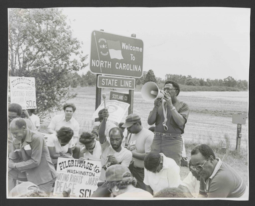 Black and white photograph of a man standing with a bullhorn, while a group of protesters sit around him.